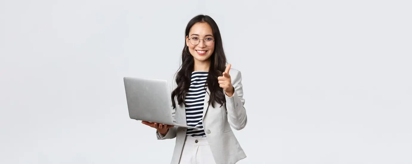 Business, finance, and employment concept featuring successful female entrepreneurs: a smiling, pleased businesswoman praising a coworker for a good point, pointing a finger at the camera, satisfied, holding a laptop.